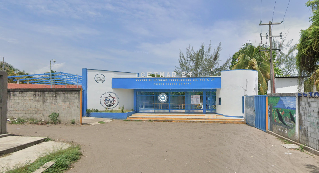 Fotografía en la que se muestra la entrada del plantel conformada por dos muros blancos y en medio de ellos una reja de color azul. El techado es de color azul y arriba hay unas letras grandes de color blanco que dicen CETMAR 24. 